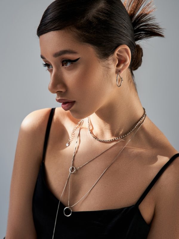 portrait of brunette asian woman with bold makeup, trendy hairstyle and expressive gaze, in black strap dress and silver necklaces looking away on grey background with lighting, trendy spring concept